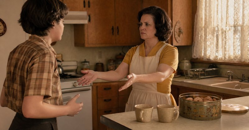 Cocina años 60: madre con delantal tendiendo los brazos con expresión tensa mientras adolescente vacila — contradicción entre gesto y afecto típica del doble vínculo